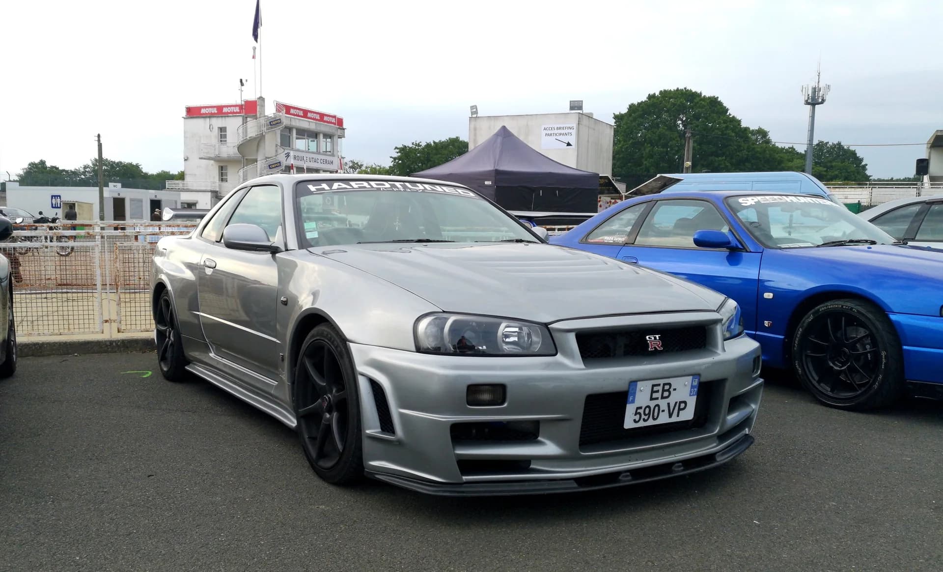 Silver Nissan Skyline GT-R R34 beside a blue coupe at a car festival.